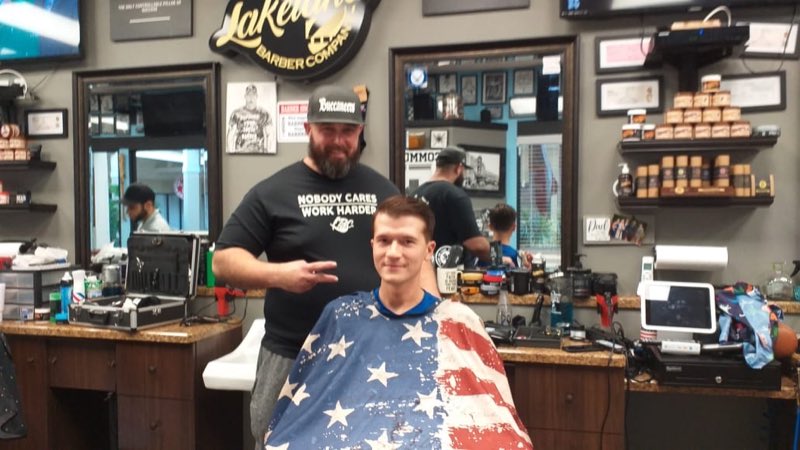 A barber posing with his young client, who wears an American-flag cape, inside the shop
